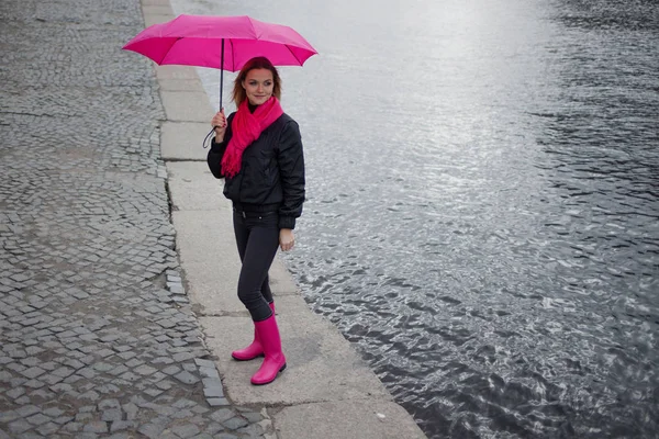 年轻和快乐金发美女在明亮的粉色围巾、 橡胶靴和起雨伞走在多雨的城市. — 图库照片