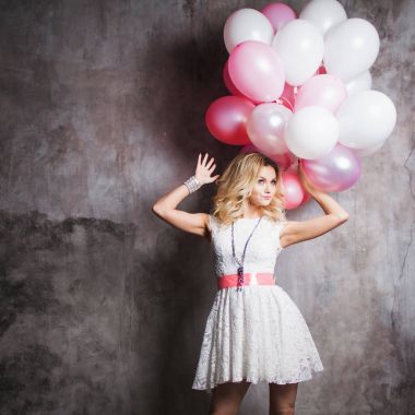 Pembe kanat ile beyaz elbiseli çekici genç sarışın balonlar büyük bir demet tutmak. Gri dokulu arka plan üzerinde