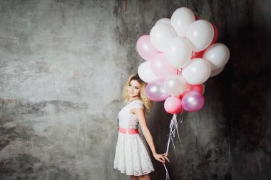 Pembe kanat ile beyaz elbiseli çekici genç sarışın balonlar büyük bir demet tutmak. Gri dokulu arka plan üzerinde