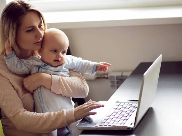 Ung mor i hjemmet kontor med computer og hendes baby. Freelancer eller blogger, arbejde og børnepasning . - Stock-foto