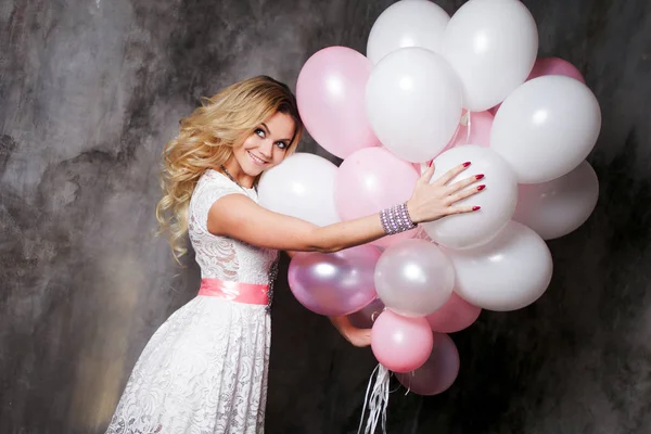Charming young blonde in a white dress with pink sash, holding a large bundle of balloons. On gray textured background