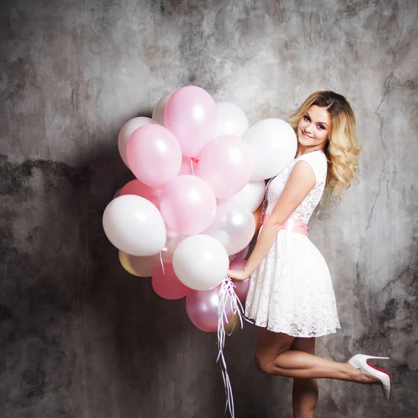 Encantadora joven rubia en un vestido blanco con faja rosa, sosteniendo un gran manojo de globos. Sobre fondo texturizado gris —  Fotos de Stock