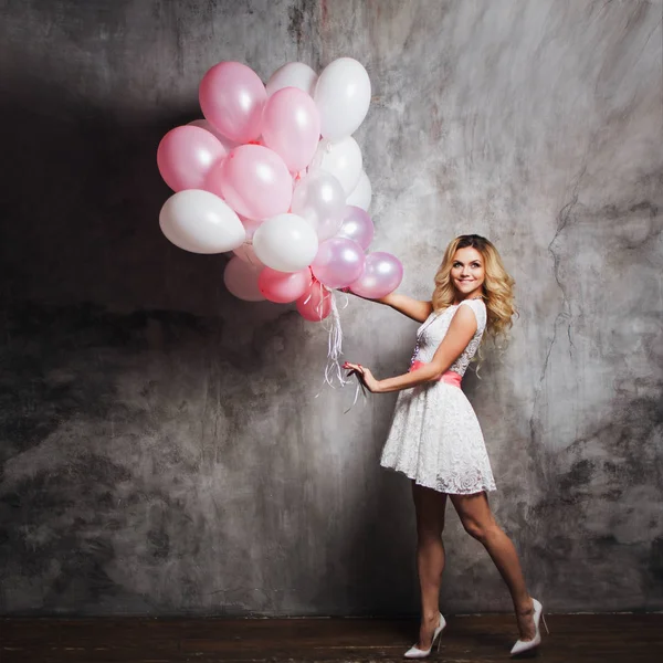 Charmante jonge blonde in een witte jurk met roze sjerp, houden een grote bundel van ballonnen. Op grijze gestructureerde achtergrond — Stockfoto