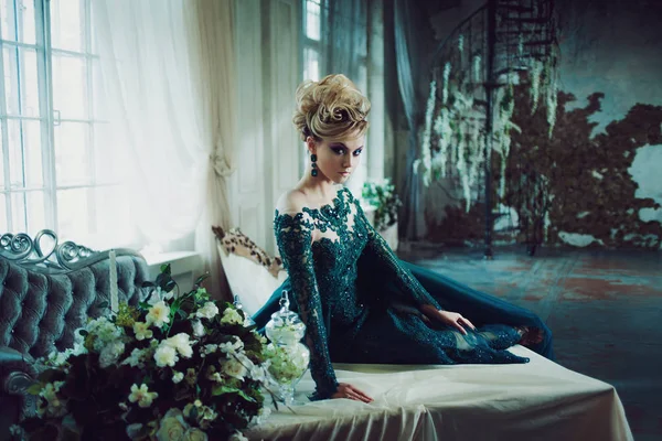 Jovem mulher loira atraente em um belo vestido verde sentado na mesa. Fundo texturizado, interior. Penteado de luxo — Fotografia de Stock
