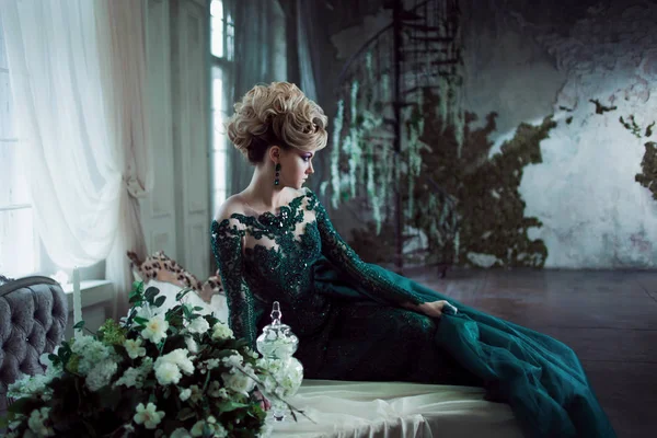 Jovem mulher loira atraente em um belo vestido verde sentado na mesa. Fundo texturizado, interior. Penteado de luxo — Fotografia de Stock