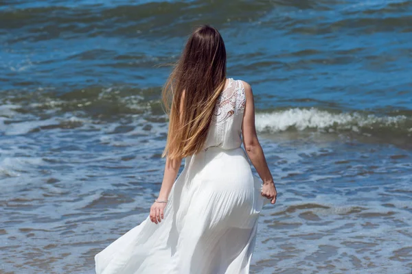 Jonge charmante brunette vrouw op Zeekust. Mooi meisje in een witte zomerjurk. Loopt naar de zee, terug weergave — Stockfoto