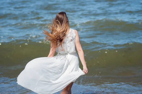 Młoda kobieta uroczy brunetka na wybrzeżu morza. Piękna dziewczyna w białych letnią sukienkę. Biegnie w kierunku morza, z powrotem widok — Zdjęcie stockowe