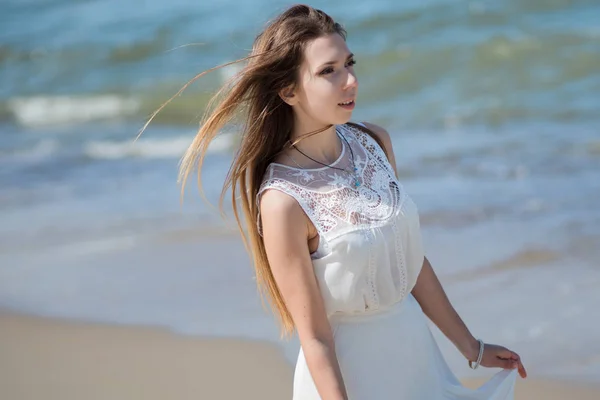 Giovane donna bruna affascinante sulla costa del mare. Bella ragazza in un abito estivo bianco. Sullo sfondo delle onde del mare — Foto Stock