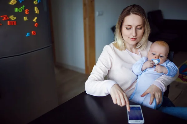 Ung mor med baby sidder og bruger telefonen. Internet browsing, indendørs - Stock-foto