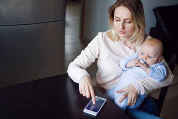 Madre joven con el bebé se sienta y utiliza el teléfono. Navegación por Internet, interior —  Fotos de Stock