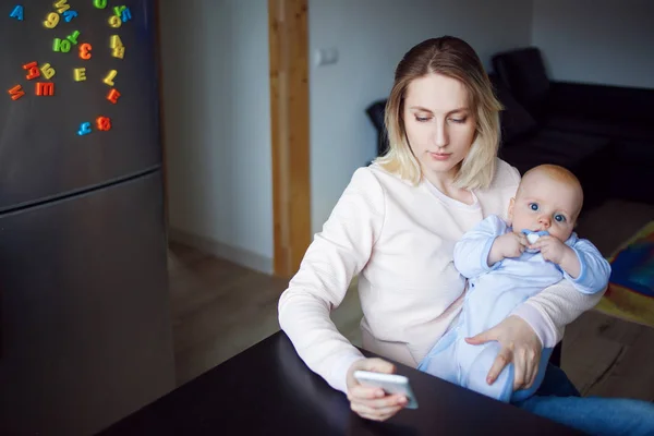 Madre joven con el bebé se sienta y utiliza el teléfono. Navegación por Internet, interior —  Fotos de Stock