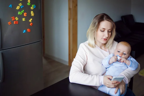 Madre joven con el bebé se sienta y utiliza el teléfono. Navegación por Internet, interior —  Fotos de Stock