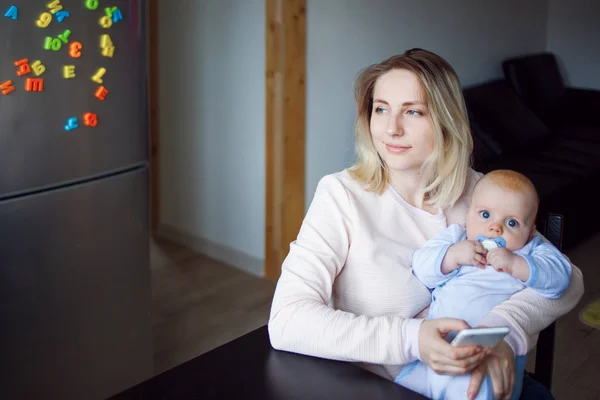 Madre joven con el bebé se sienta y utiliza el teléfono. Navegación por Internet, interior —  Fotos de Stock