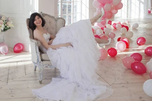 Mujer joven en vestido de novia en el interior de lujo con una masa de globos rosados y blancos, sentado en la silla. Encantadora joven novia morena con corte de pelo corto en vestidos de Quinceañera con estilo . —  Fotos de Stock