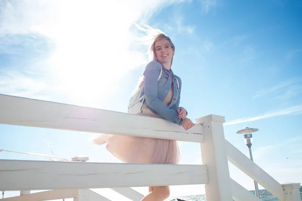 Vrolijk meisje aan de kust leunend op de houten witte hek. Jonge blonde vrouw die lacht. Trendy beige rok — Stockfoto