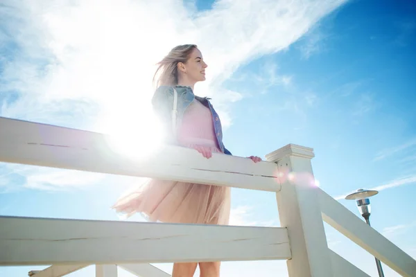 Joven chica alegre en la orilla del mar apoyada sobre la cerca blanca de madera. Mujer rubia joven sonriendo. Falda beige de moda —  Fotos de Stock