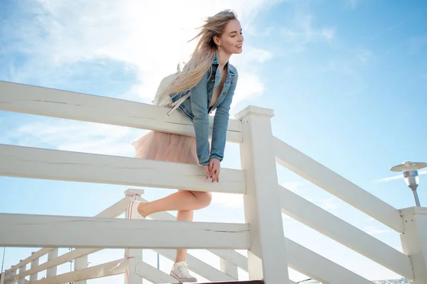 Junges fröhliches Mädchen am Ufer des Meeres, das sich über den hölzernen, weißen Zaun beugt. junge blonde Frau lächelnd. trendiger beiger Rock — Stockfoto