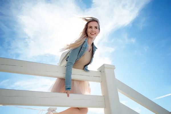 Jovem menina alegre na praia inclinando-se sobre a cerca branca de madeira. Jovem loira sorrindo. Saia bege na moda — Fotografia de Stock