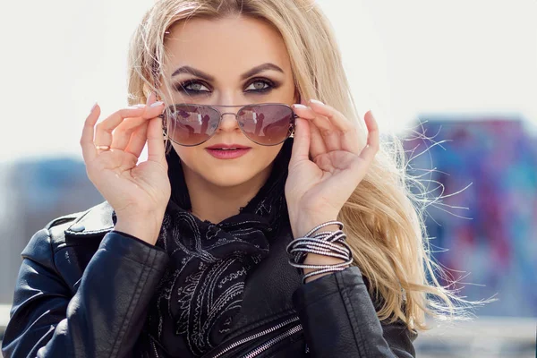 Close up image of beautiful young woman looking at camera. Young caucasian female model looking through sunglasses. Femme blonde in a leather jacket — Stock Photo, Image
