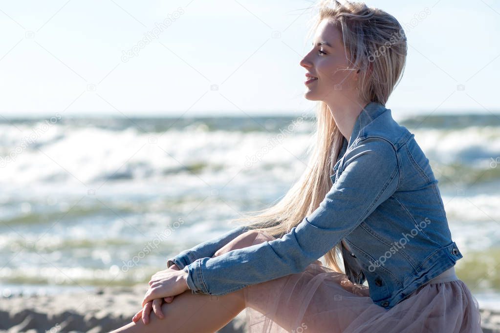 Young cheerful girl on the seashore. Young blonde woman sitting on the sand. Trendy beige skirt