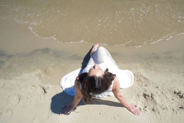 Jonge mooie brunette vrouw in witte jurk aan de kust, zittend op het zand. Bovenaanzicht — Stockfoto