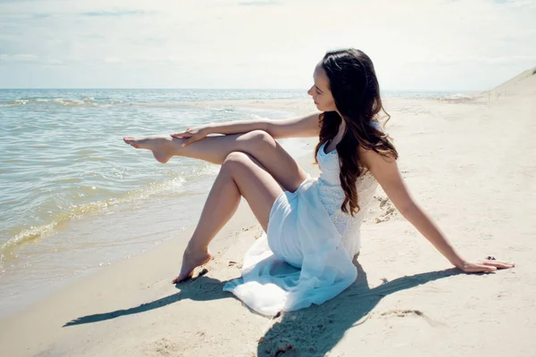 Όμορφη μελαχρινή νεαρή γυναίκα σε λευκό φόρεμα στην παραλία. Χαϊδεύοντας ένα πόδι με το ένα χέρι — Φωτογραφία Αρχείου