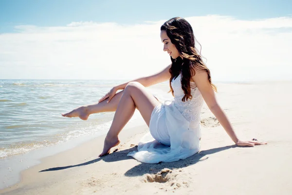 Young beautiful brunette woman in white dress on the seashore. Sitting on the sand — Stock Photo, Image