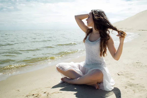 Jonge mooie brunette vrouw in witte jurk aan de kust, zittend op het zand en kijkt naar de horizon — Stockfoto