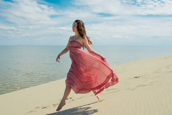 Kvinna i rött viftande klänning med flygande tyg körs på bakgrund av sanddyner. Skyline och havet — Stockfoto