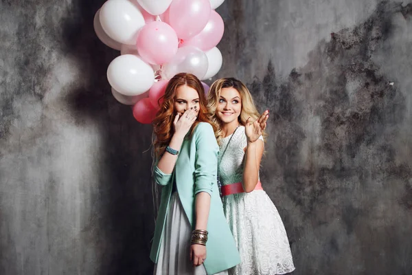 Twee meisjes lachen en kijken naar de zijde, te bespreken iemand. Jonge charmante vriendinnen op het feest. Gelukkig en vrolijk meisje met ballonnen. Op grijze gestructureerde achtergrond — Stockfoto