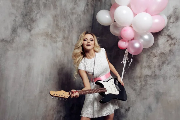 Adorable young blonde with guitar at the party. Holding a large bundle of balloons. On gray textured background.