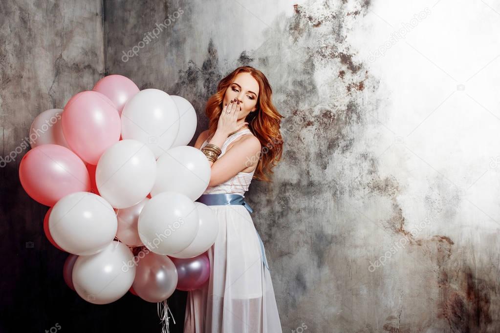 Red-haired beauty young woman in a white dress, holding a large bundle of balloons at the party.