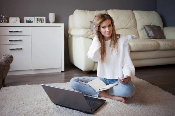 Charmantes junges Mädchen in weißem Pullover und Jeans sitzt mit Notizbuch auf dem Boden. — Stockfoto