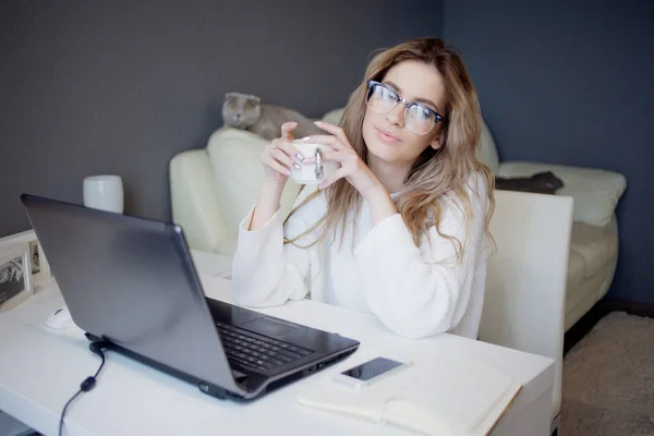 Φοιτητή ή ελεύθερος επαγγελματίας, εργάζεται στο σπίτι με το laptop. Γοητευτική νεαρή γυναίκα κάθεται μπροστά από την οθόνη με καφέ. — Φωτογραφία Αρχείου