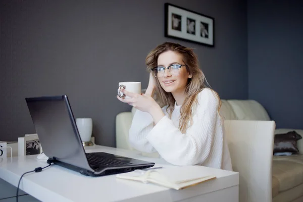 Estudiante o freelancer, trabajando en casa con laptop. Encantadora joven se sienta frente al monitor con una taza de café . — Foto de Stock