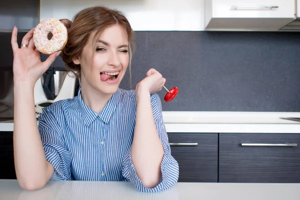 Beautiful young girl with a donut and a Lollipop in the kitchen. To do funny faces — Stock Photo, Image