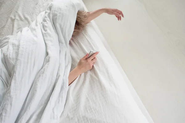 De vrouw onder de deken zet de wekker uit of kijkt naar het sociale netwerk. — Stockfoto