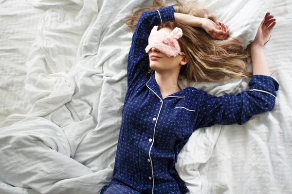 Niedliche Blondine in ihrem Bett im blauen Pyjama und Schlafmaske, Draufsicht — Stockfoto