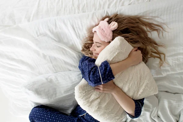 Linda rubia en su cama en pijama azul y máscara del sueño, almohada abrazo — Foto de Stock
