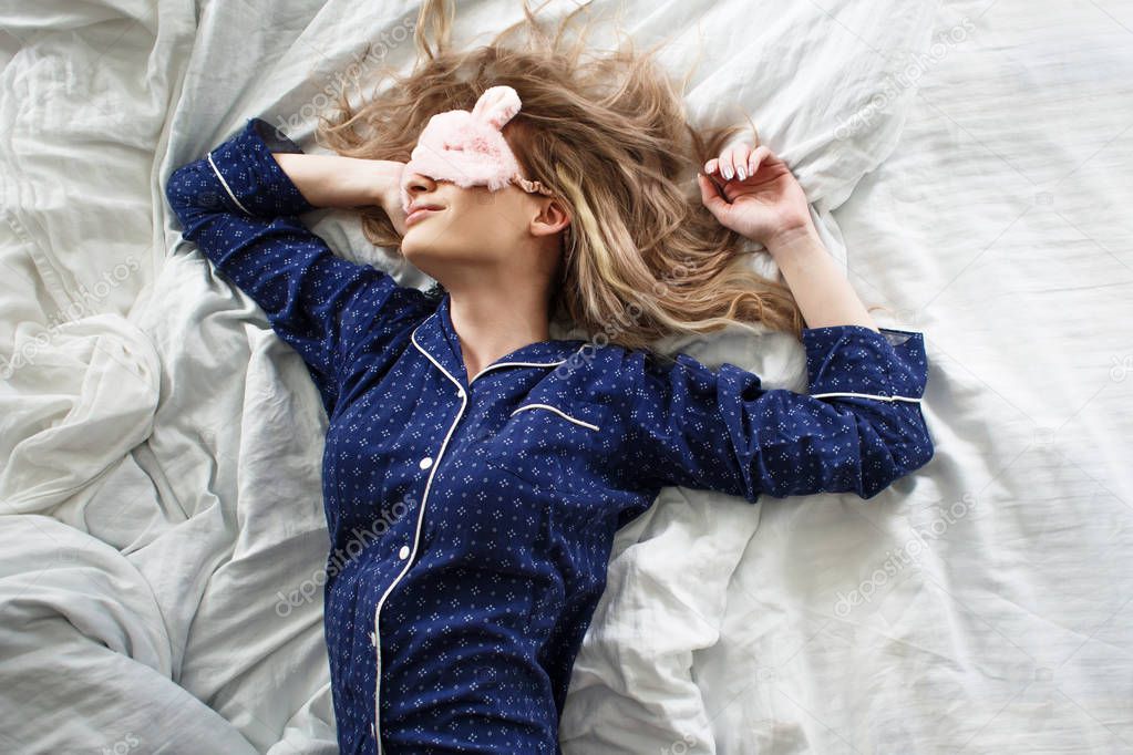 Cute blonde in her bed in blue pajamas and sleep mask, top view