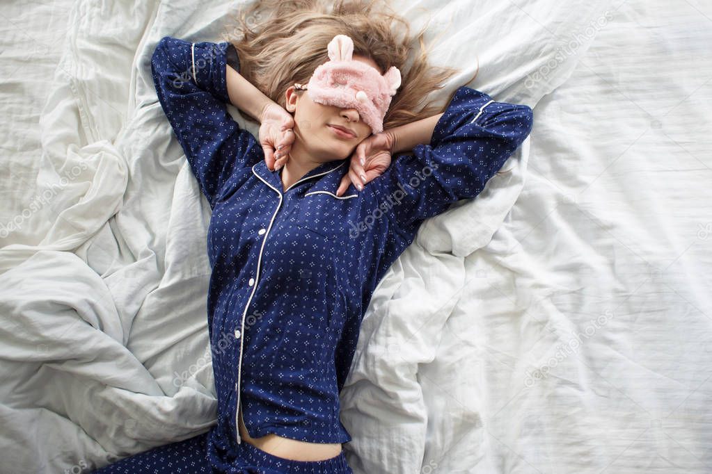 Cute blonde in her bed in blue pajamas and sleep mask, top view