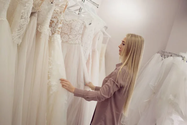Jeune femme blonde ressemble robe différente sur le cintre. Boutique de mariée . — Photo