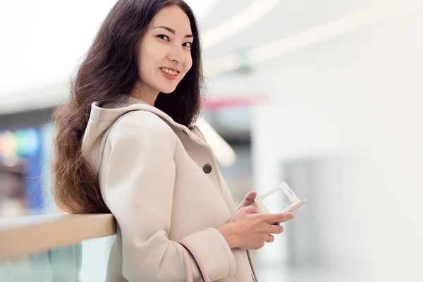 Young beautiful girl Asian, uses mobile phone, background, shopping Mall or business center — Stock Photo, Image