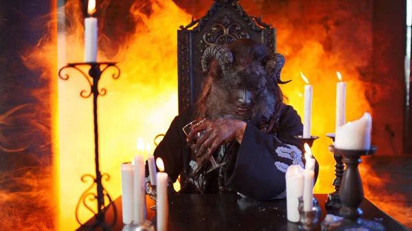 Homem na imagem de monstro com chifres sentado à mesa com um copo na mão . — Fotografia de Stock