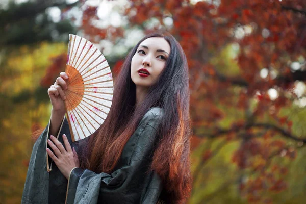 Beautiful young Asian woman with fan on background of red maple — Stock Photo, Image