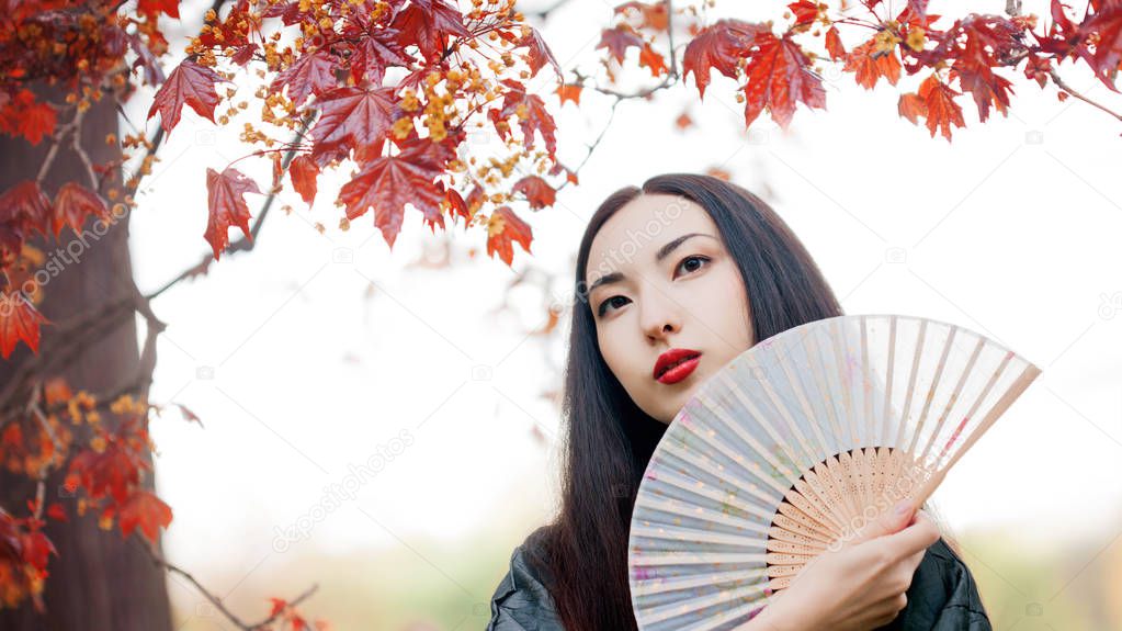 Beautiful young Asian woman with fan on background of red maple