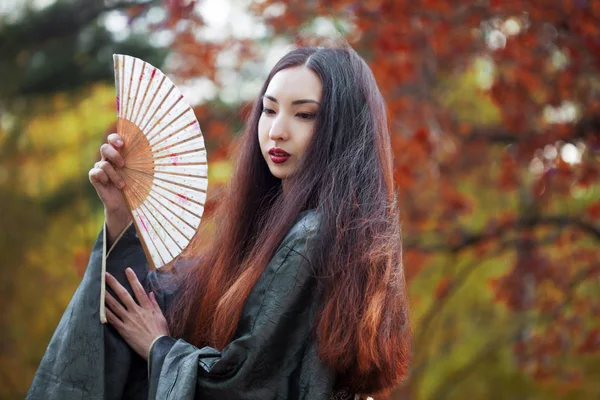 Beautiful young Asian woman with fan on background of red maple — Stock Photo, Image