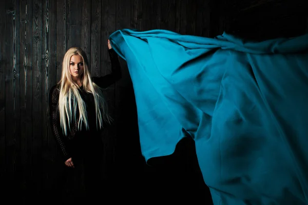 Snygg blondin på en mörk bakgrund med ljusa flygande tyg, färg blå — Stockfoto
