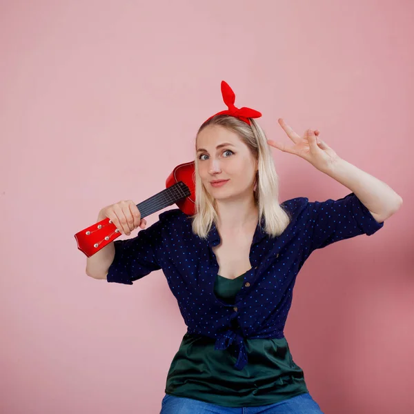 Portret van stijlvolle jonge blonde met ukulele. Een vrouw met een hoofdband op een roze achtergrond — Stockfoto