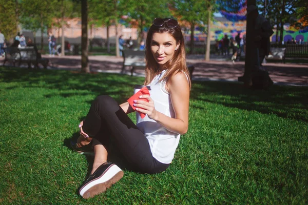 Молода красива жінка сидить на траві і п'є каву, щоб піти — стокове фото